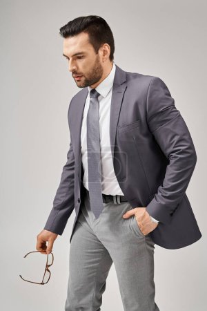 hombre de negocios equilibrado en ropa formal sosteniendo gafas y caminando con la mano en el bolsillo sobre fondo gris