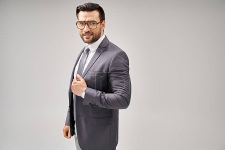cheerful businessman in formal wear and glasses standing on grey background, elegance and style