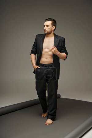 fashion statement, barefoot and shirtless man in pinstripe suit posing on grey background