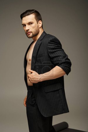 Mode-Statement, gutaussehender und hemdsloser Mann im Nadelstreifenanzug posiert auf grauem Hintergrund