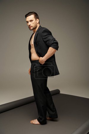 fashionable and handsome man with bare chest and pinstripe suit posing on grey background