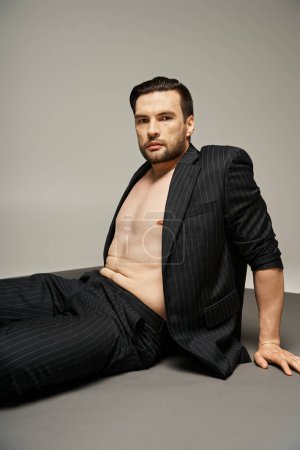 Porträt eines eleganten und gut aussehenden Mannes mit nackter Brust, der in Nadelstreifengewand auf grauem Hintergrund posiert