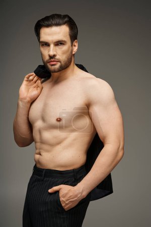 Shirtloser und gutaussehender Mann mit Borsten und nackter Brust posiert in Nadelstreifenhosen und Halsjacke