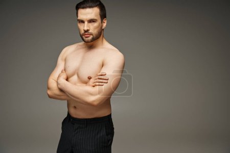 Porträt eines brünetten und gut aussehenden Mannes mit nackter Brust, der mit verschränkten Armen auf grauem Hintergrund posiert