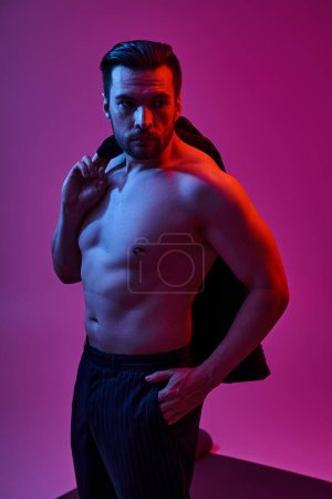 seductive shirtless man with bare chest and bristle holding jacket while posing on purple backdrop