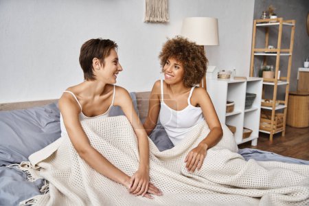 Téléchargez les photos : Femme américaine africaine joyeuse avec les cheveux bouclés assis avec petite amie sur le lit le matin, confortable - en image libre de droit