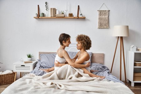 cheerful multicultural and young lesbian couple hugging and looking at each other in bedroom