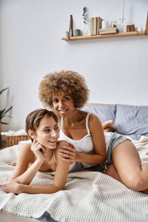 Téléchargez les photos : Joyeuse femme afro-américaine étreignant petite amie tout en étant couché ensemble sur le lit, l'intimité et l'amour - en image libre de droit