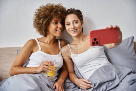 glückliche Frau macht Selfie auf Smartphone nahe afrikanisch-amerikanischer Freundin mit einem Glas Orangensaft