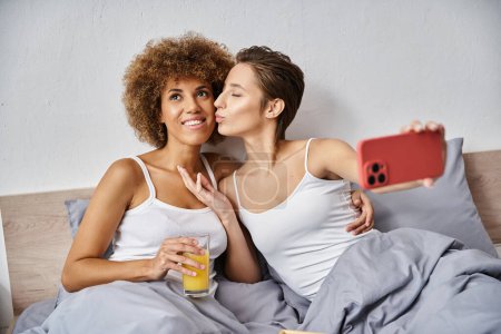 glückliche Frau macht Selfie, während sie afrikanische amerikanische Freundin mit einem Glas Orangensaft küsst