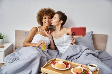 happy woman taking selfie while kissing african american girlfriend near breakfast tray on bed