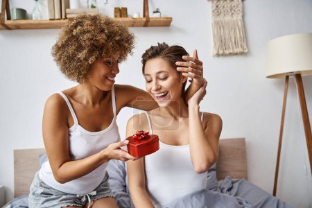african american woman holding red gift near excited girlfriend in pajamas on valentines day
