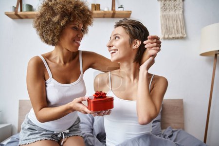 joyful african american woman holding red gift near excited girlfriend in pajamas on valentines day