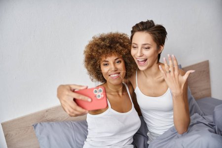 excited and engaged interracial lesbian couple taking selfie on smartphone at home, engagement ring