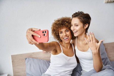 happy and engaged interracial lesbian couple taking selfie on smartphone at home, engagement ring
