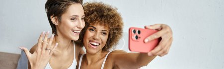 happy and engaged interracial lesbian couple taking selfie on smartphone at home, banner