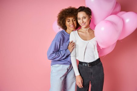 Valentines day concept, joyful multicultural lesbian couple smiling near heart shaped balloons