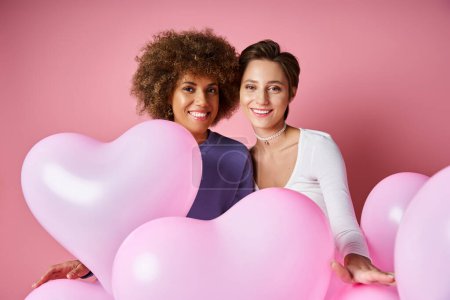 Valentines day concept, happy multicultural lesbian couple smiling near pink heart shaped balloons