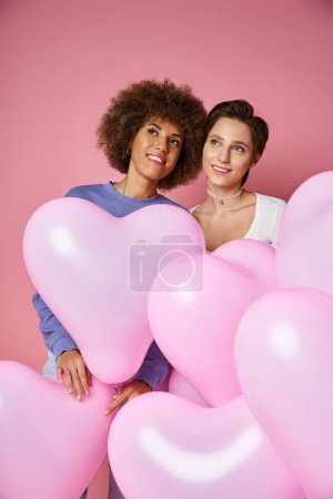 verträumtes lesbisches Paar lächelt in der Nähe rosafarbener Herzballons, Valentinstag