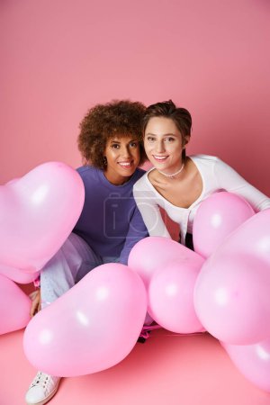 glückliches multikulturelles lesbisches Paar, das zusammen in der Nähe rosa herzförmiger Luftballons sitzt, Valentinstag