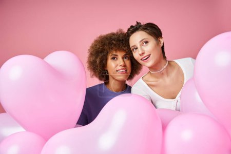 dreamy multicultural lesbian couple looking away near pink heart shaped balloons, Valentines day