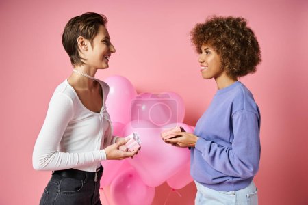 happy multicultural lesbian couple exchanging gifts near heart shaped balloons on Valentines day
