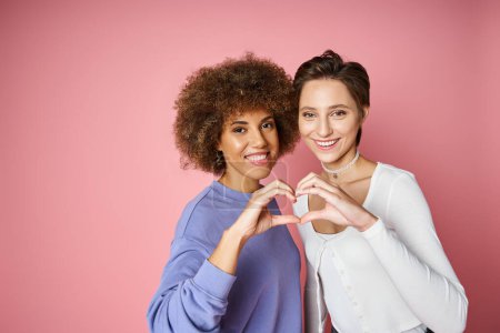 fröhliches multikulturelles und lesbisches Paar zeigt Herzzeichen mit Händen auf rosa Hintergrund, lgbtq love