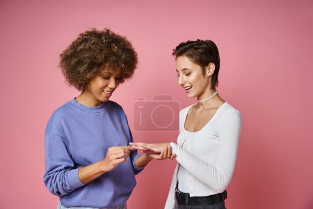 afrikanisch-amerikanische Frau trägt Verlobungsring am Finger der erstaunten Freundin auf rosa Hintergrund