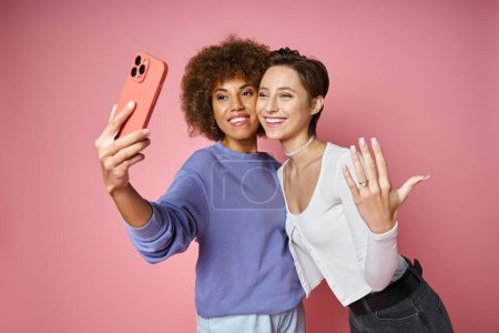 happy lesbian woman showing engagement ring on her finger while taking selfie with fiancee on pink