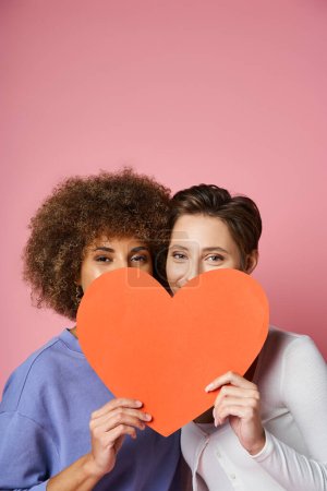 happy lesbian and multiethnic couple hiding behind red carton heart on pink, Valentines day