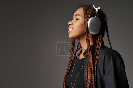pleased african american girl in 20s with dreadlocks listening music in headphones on grey backdrop