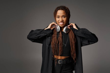 smiling african american woman with dreadlocks standing in headphones and looking at camera on gray