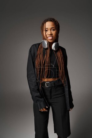 happy african american woman with dreadlocks standing in headphones and looking at camera on gray