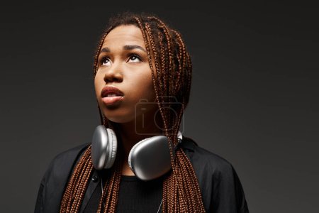 dreamy african american woman with dreadlocks standing in headphones and looking up on grey