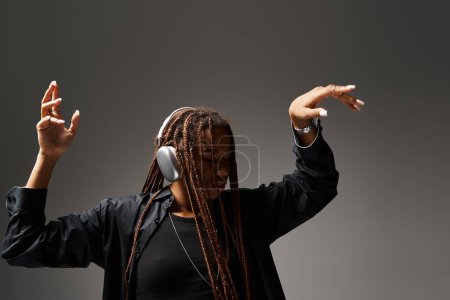 ecstatic dance of african american girl in her 20s with dreadlocks in wireless headphones on grey