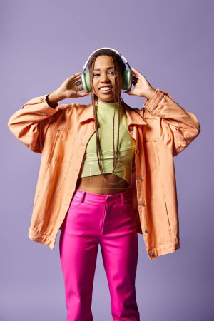 Téléchargez les photos : Heureuse fille afro-américaine dans des écouteurs sans fil et tenue décontractée dynamique sur fond violet - en image libre de droit