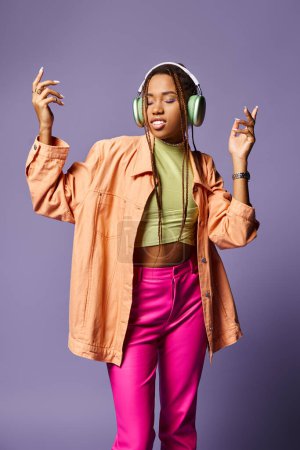 danza extática de la muchacha afroamericana feliz en traje vibrante con auriculares en el telón de fondo púrpura