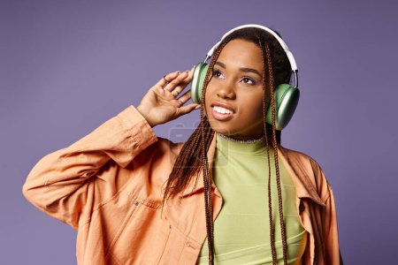 Téléchargez les photos : Heureuse femme afro-américaine en tenue tendance vibrante avec écouteurs sans fil sur fond violet - en image libre de droit