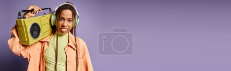 Téléchargez les photos : Heureuse femme afro-américaine dans des écouteurs sans fil debout avec boombox rétro sur violet, bannière - en image libre de droit