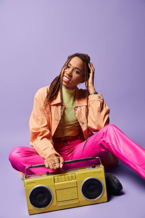 Happy african american woman with dreadlocks sitting next to retro boombox on purple backdrop