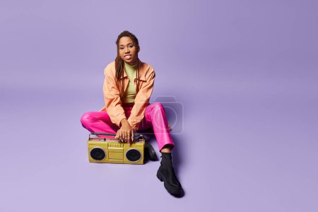 Téléchargez les photos : Femme américaine africaine joyeuse avec dreadlocks assis à côté de boombox rétro sur fond violet - en image libre de droit