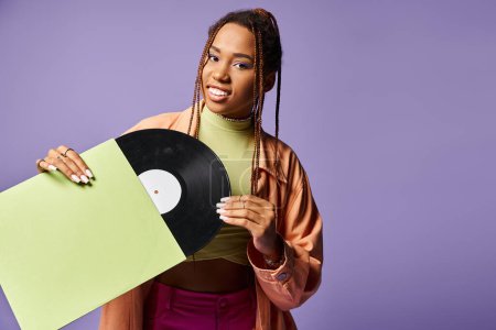 Téléchargez les photos : Heureuse jeune femme afro-américaine dans la vingtaine avec dreadlocks tenant disque de vinyle sur fond violet - en image libre de droit