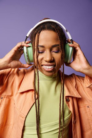 joyful african american woman in vibrant attire with wireless headphones on purple backdrop