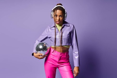 young african american woman in trendy attire and headphones holding disco ball on purple backdrop