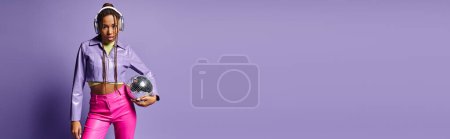 Téléchargez les photos : Jeune femme afro-américaine en tenue tendance et écouteurs tenant boule disco sur violet, bannière - en image libre de droit