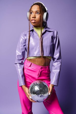 Foto de Mujer afroamericana joven en auriculares inalámbricos de moda sosteniendo bola disco sobre fondo púrpura - Imagen libre de derechos