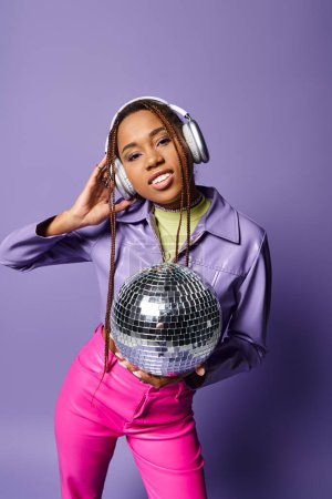 happy young african american woman in stylish attire and wireless headphones holding disco ball