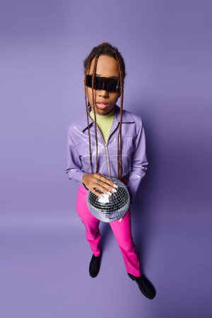 Photo for Young african american woman in trendy sunglasses holding disco ball on chain and standing on purple - Royalty Free Image