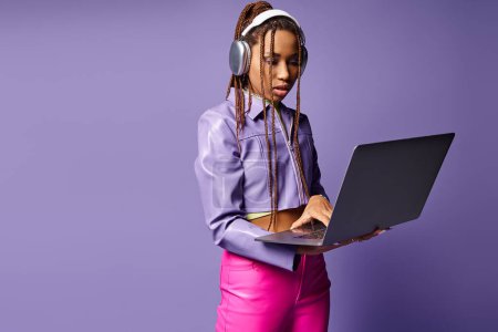 Focused african american woman with headphones working on laptop remotely on purple background