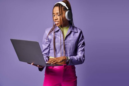 happy african american woman with headphones working on laptop remotely on purple background